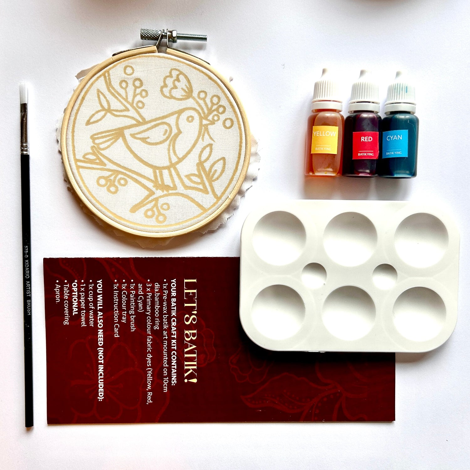 Embroidery kit laid out on a white background, including a wooden hoop with a bird embroidery design, a paintbrush, three small bottles of thread or ink in amber, red, and blue, a white six-compartment palette, and a dark red instruction card.