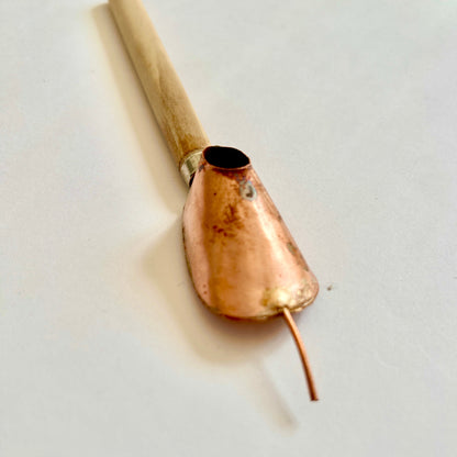 A traditional batik tjanting tool with a wooden handle and copper wax reservoir, featuring a fine spout for applying hot wax onto fabric.