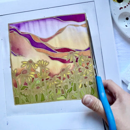 Hand holding a blue tool over a painted landscape with mountains and flowers on a white surface.